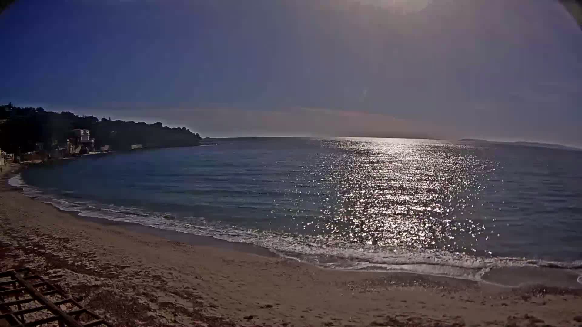 A sandy beach meets a calm, sunlit sea under a clear blue sky, with a distant shoreline visible.