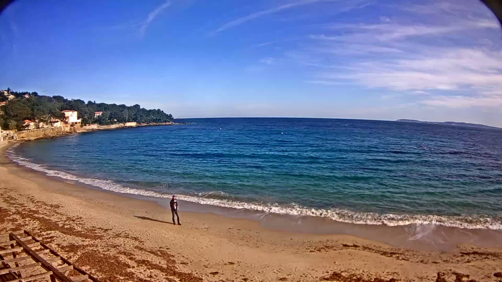 A person stands on a sandy beach next to a calm, blue ocean under a mostly sunny sky.