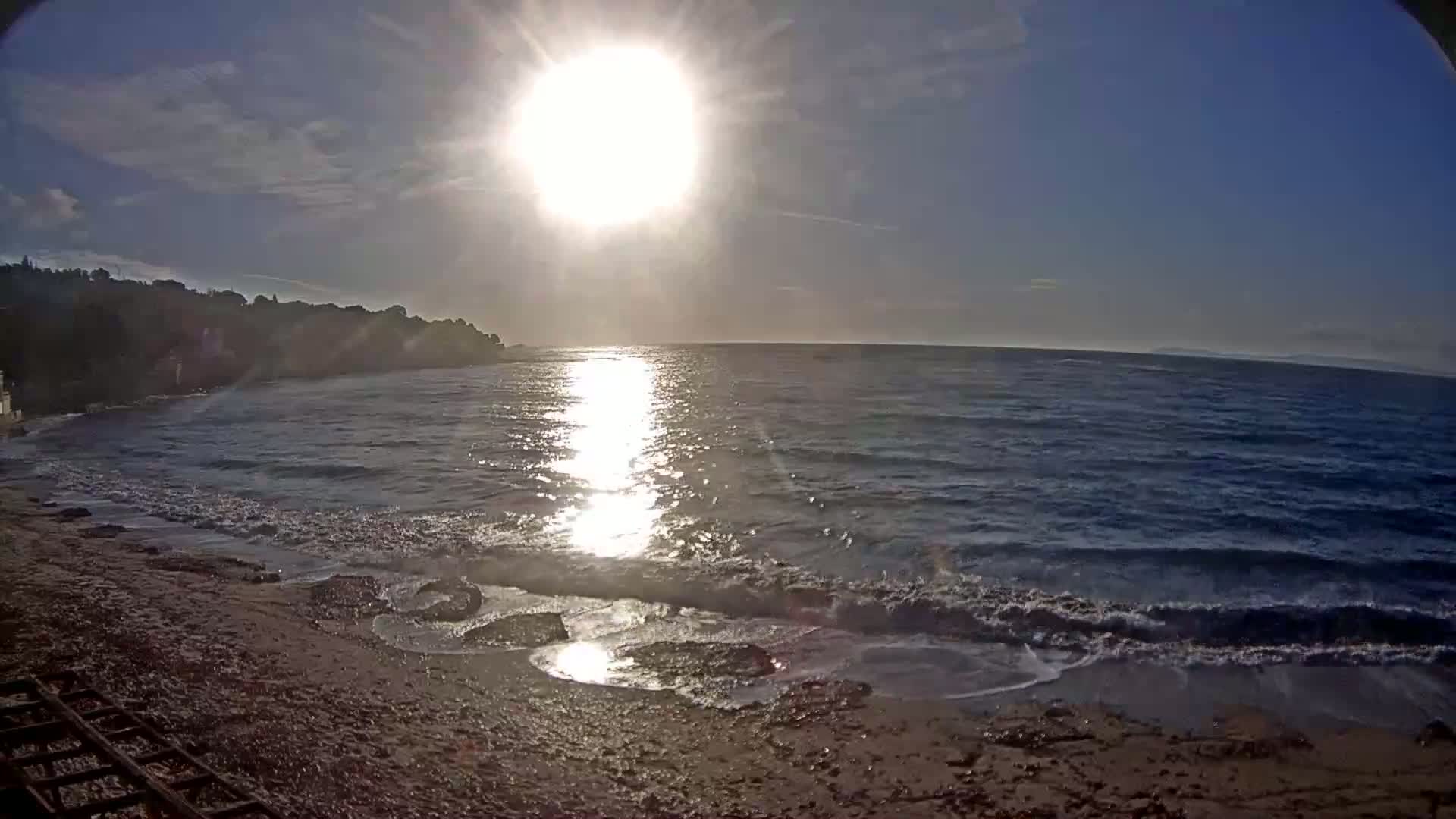The bright sun shines over a calm sea with small waves lapping a sandy beach under a mostly clear sky.