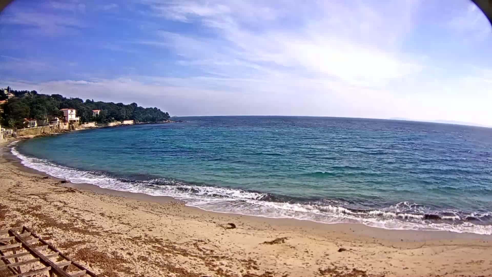 A sandy beach with small waves laps the shore, under a mostly sunny sky, next to a calm sea with houses visible on a hill in the background.