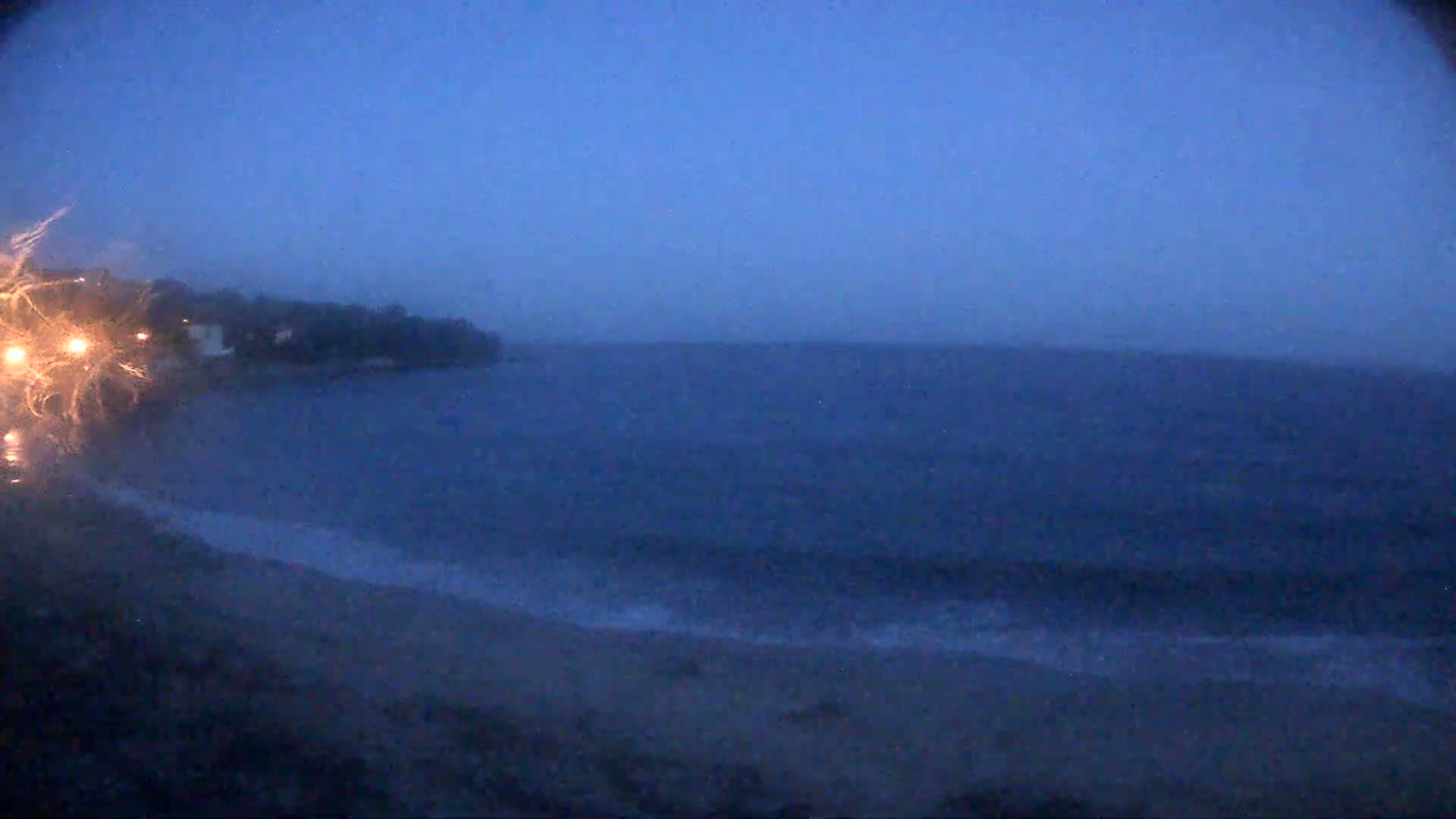 A dark, stormy ocean meets a shoreline at dusk, with headlights visible in the foreground.