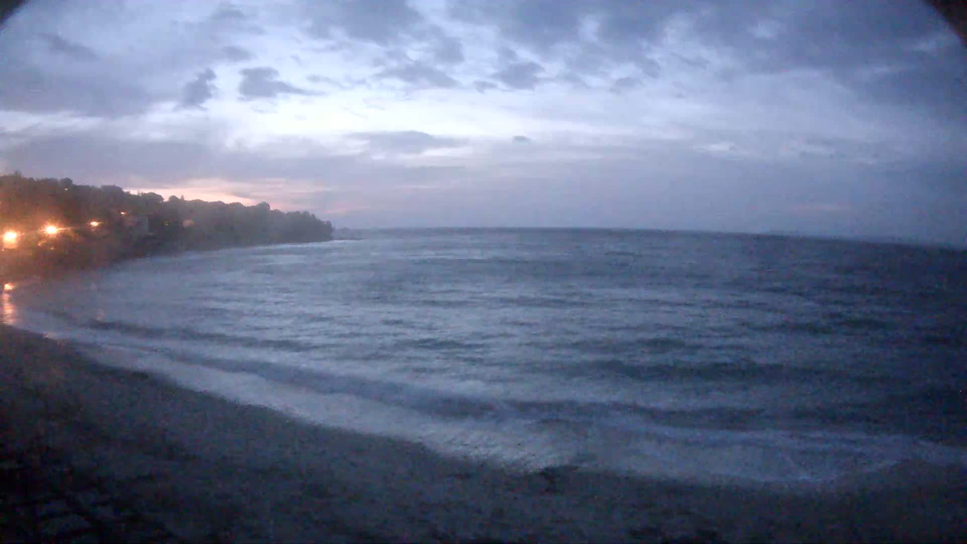 A dark, overcast sky hangs over a beach with gently lapping waves, and a dimly lit shoreline visible in the distance.