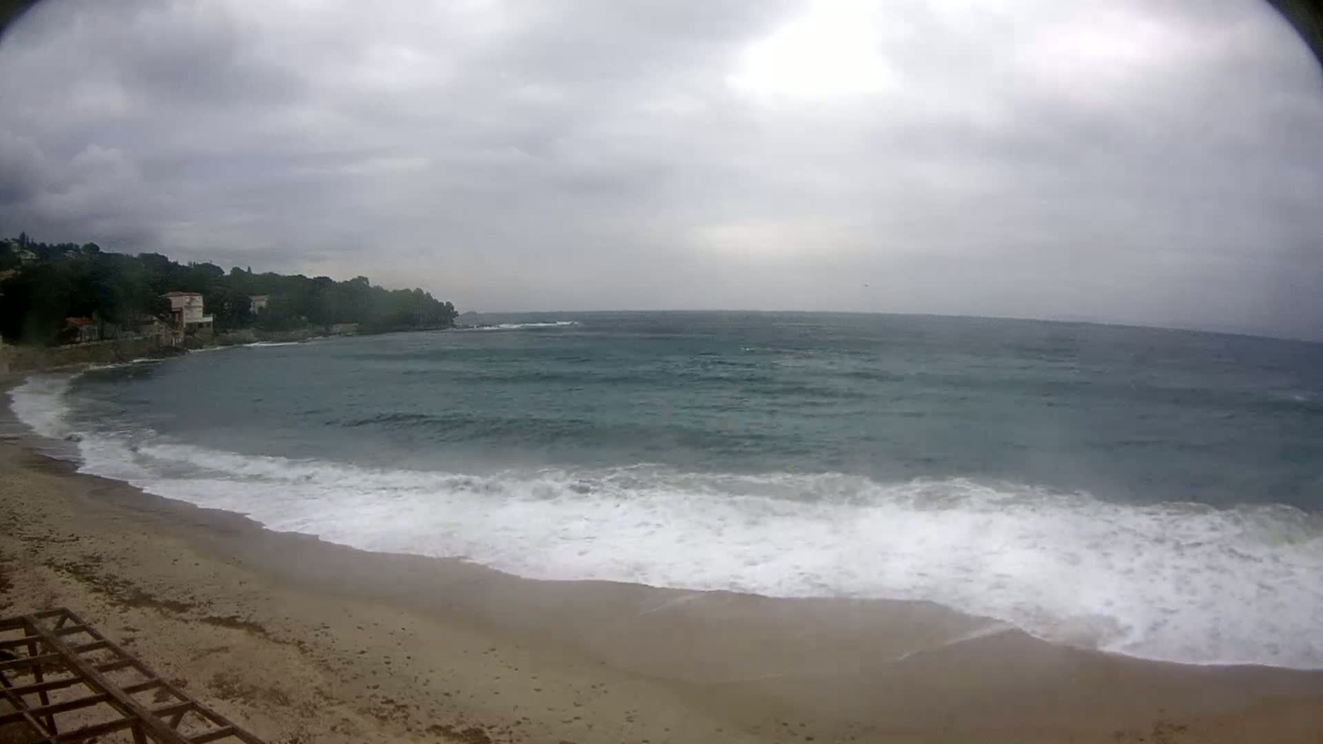 A sandy beach with moderate waves rolling onto the shore under an overcast sky, backed by a line of trees and buildings.