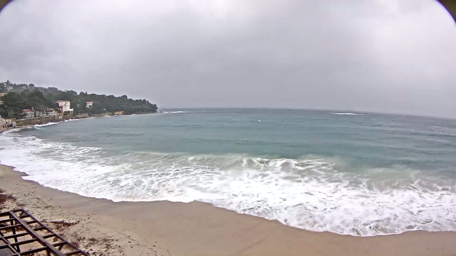 Waves crash on a sandy beach near a line of houses nestled amongst trees on a cloudy day.