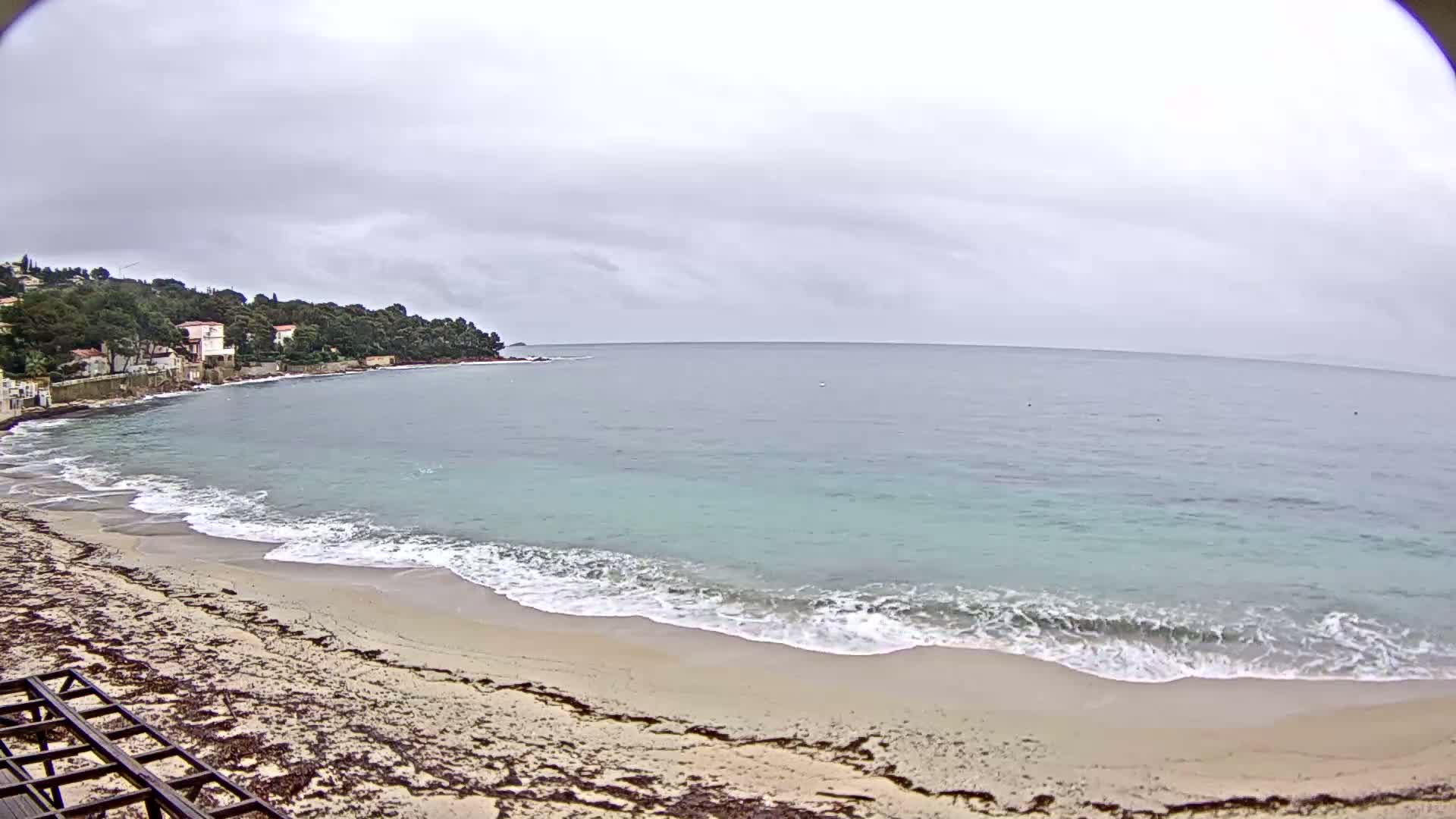 A sandy beach with gentle waves laps the shore under an overcast sky, with houses visible on a tree-lined hill in the background.