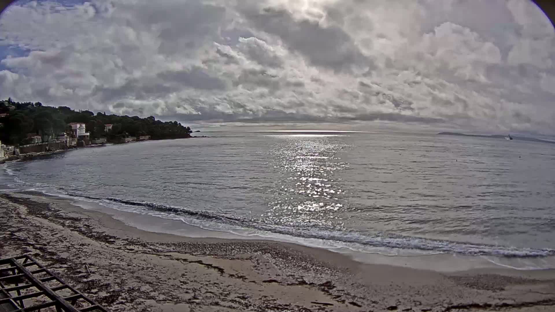 A sandy beach with gentle waves laps the shore under a partly cloudy sky, with houses and trees visible on a hillside in the background.