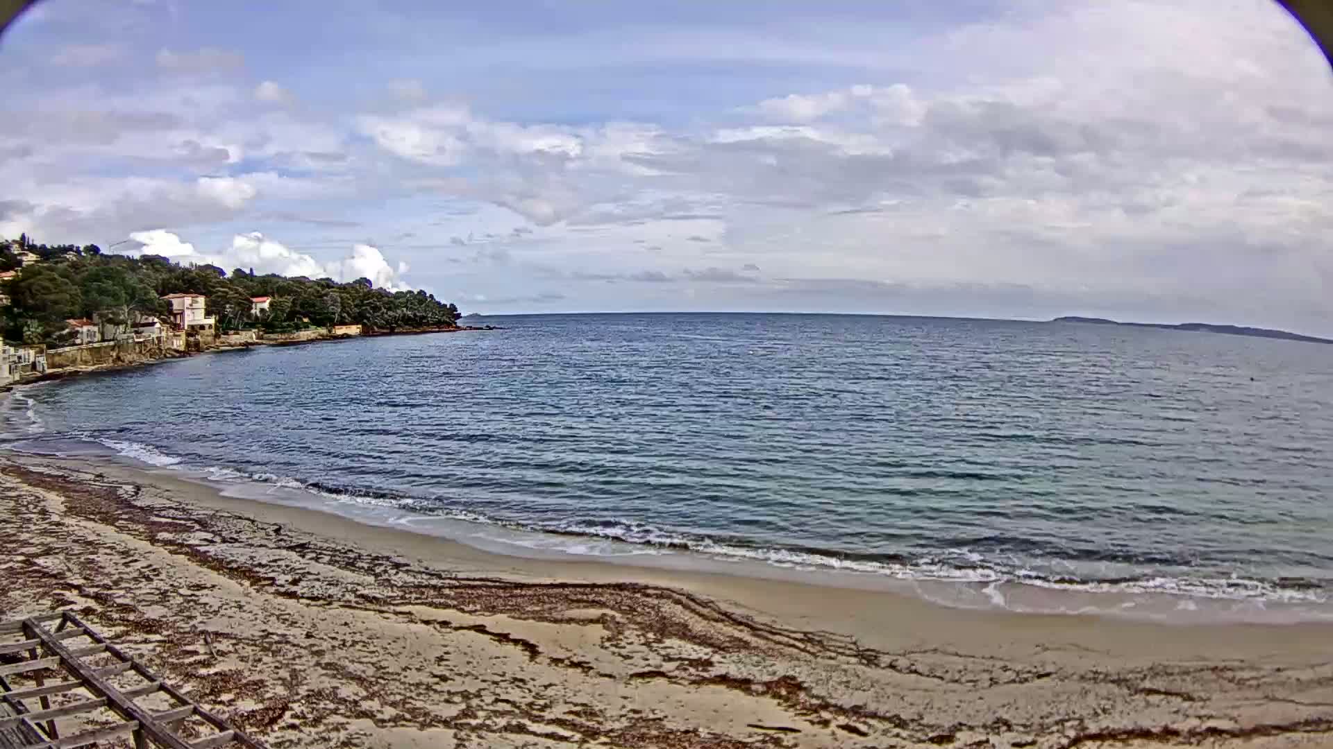 A sandy beach with small waves laps the shore under a partly cloudy sky, with houses and trees visible on a nearby hillside.