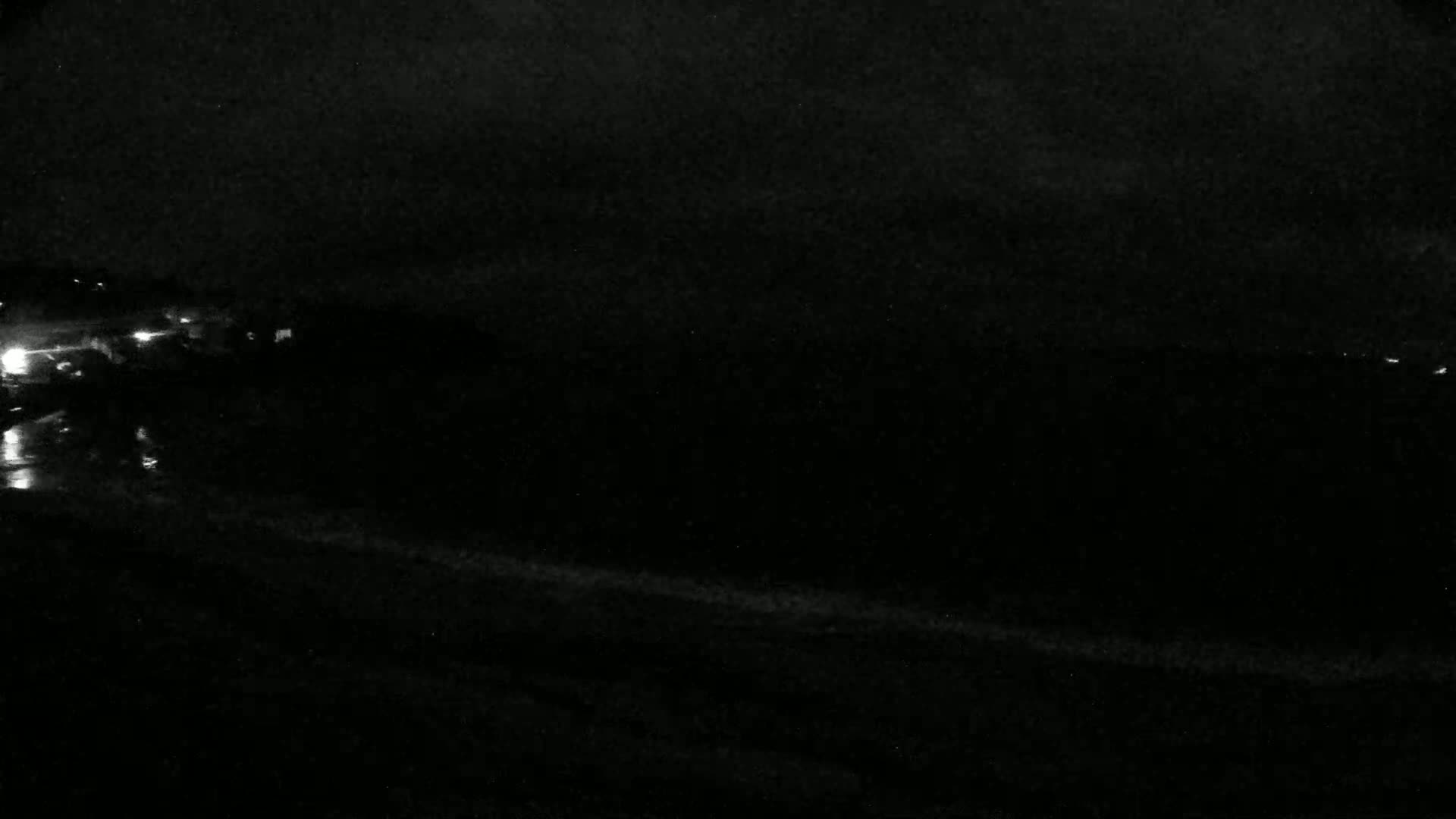 A dark nighttime scene shows a body of water under a mostly cloudy sky, with faint lights visible in the distance on land.