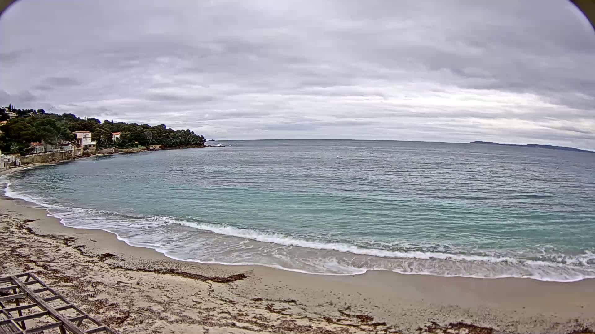 A sandy beach with gentle waves laps the shore under a cloudy sky, with houses nestled on a tree-covered hillside in the background.