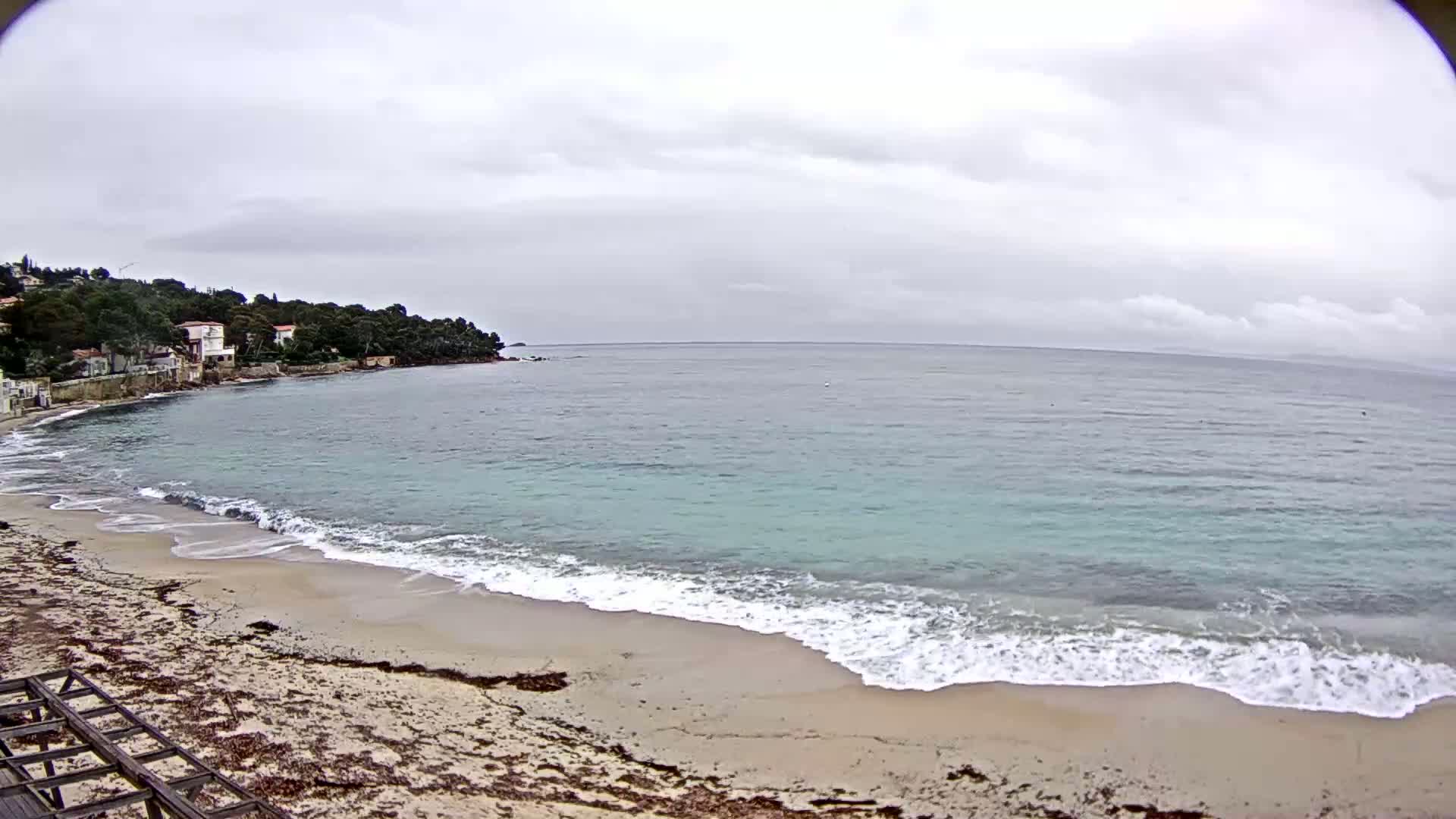 A sandy beach with small waves laps the shore under a cloudy sky, with houses and trees visible on a nearby hillside.