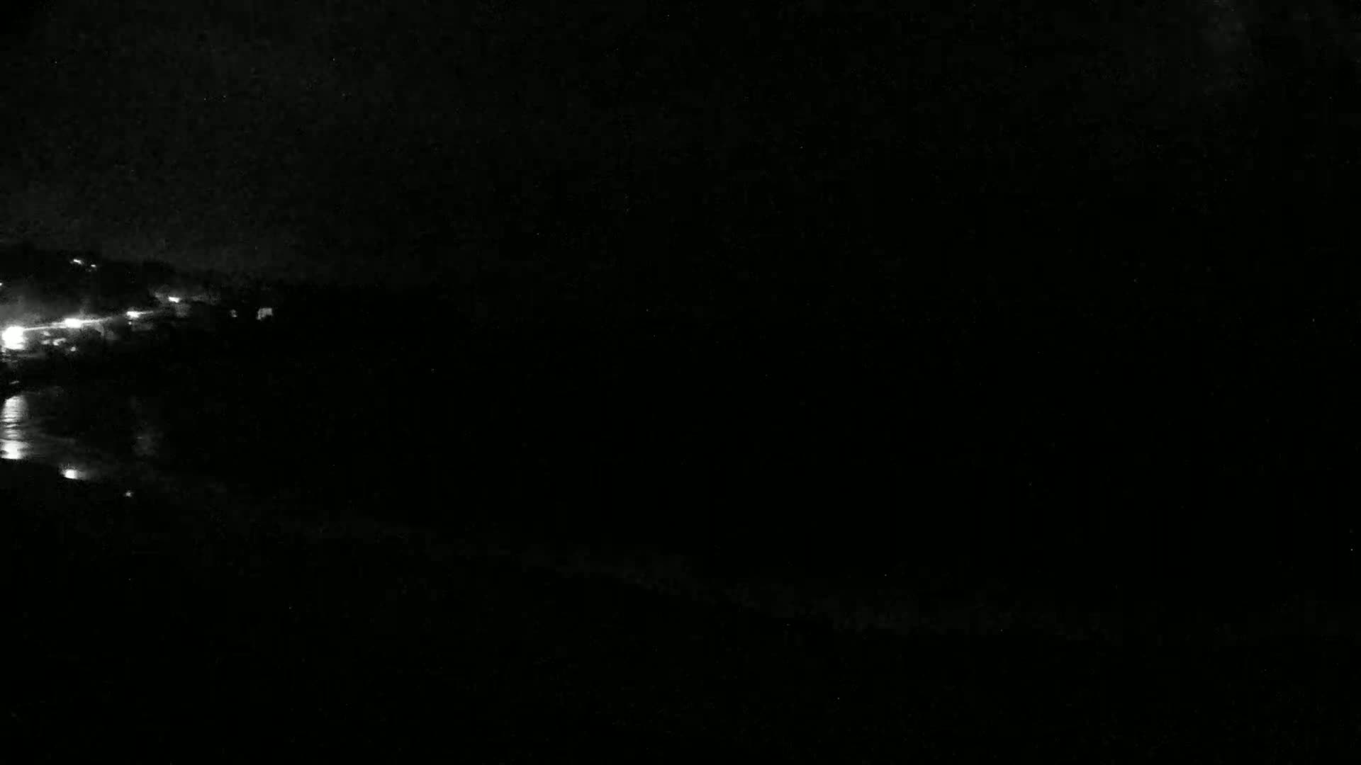 A dark, nighttime scene shows a body of water reflecting faint lights from a distant shoreline under a mostly clear sky.