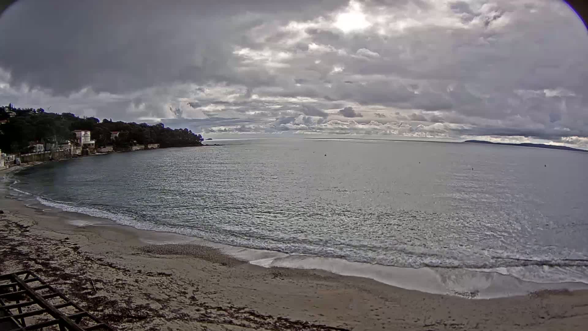 A sandy beach with gentle waves laps the shore under a cloudy sky, with houses visible on a hillside across a calm bay.