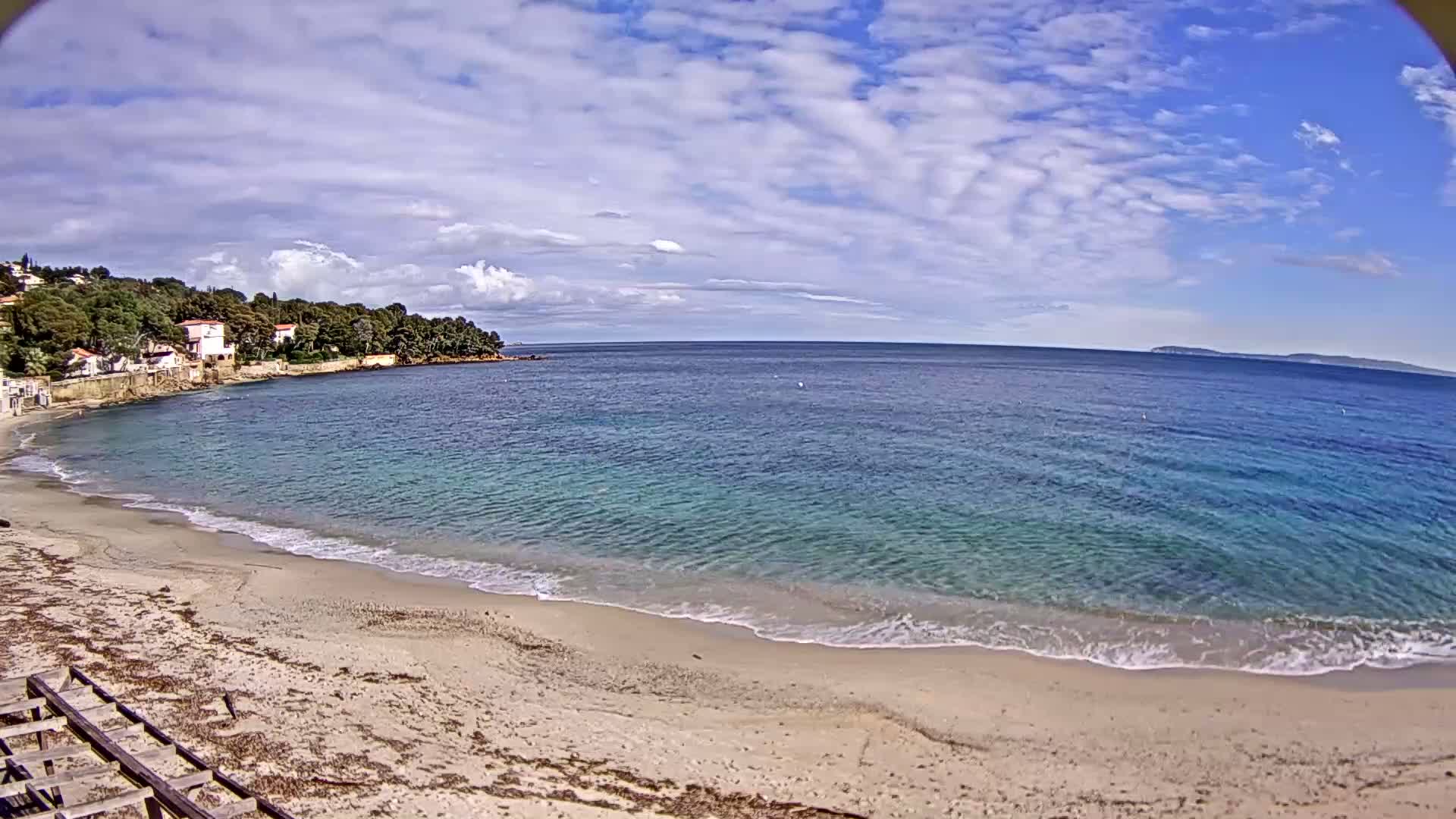 A sandy beach with gentle waves laps the shore under a partly cloudy sky, backed by a line of houses nestled amongst trees.