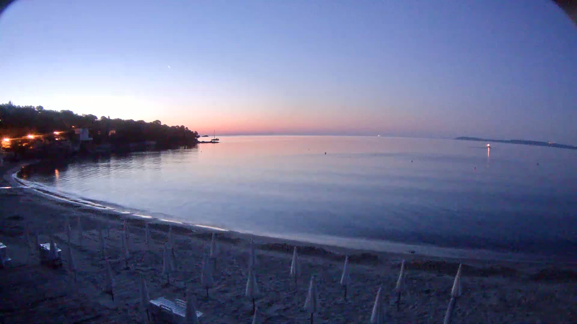 A calm, twilight-lit ocean bay is seen from a sandy beach lined with closed umbrellas, with a dark shoreline and buildings visible in the distance.
