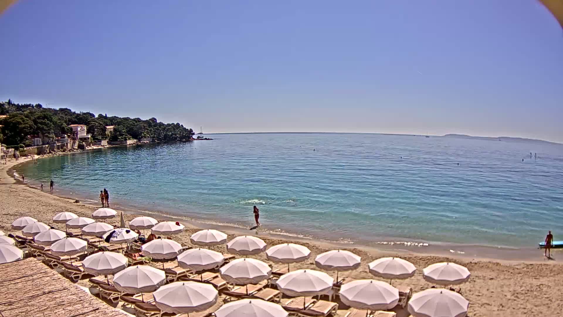 A sandy beach with rows of white umbrellas lines a calm, clear turquoise sea under a bright, sunny sky, backed by a tranquil waterfront town nestled among green hills.