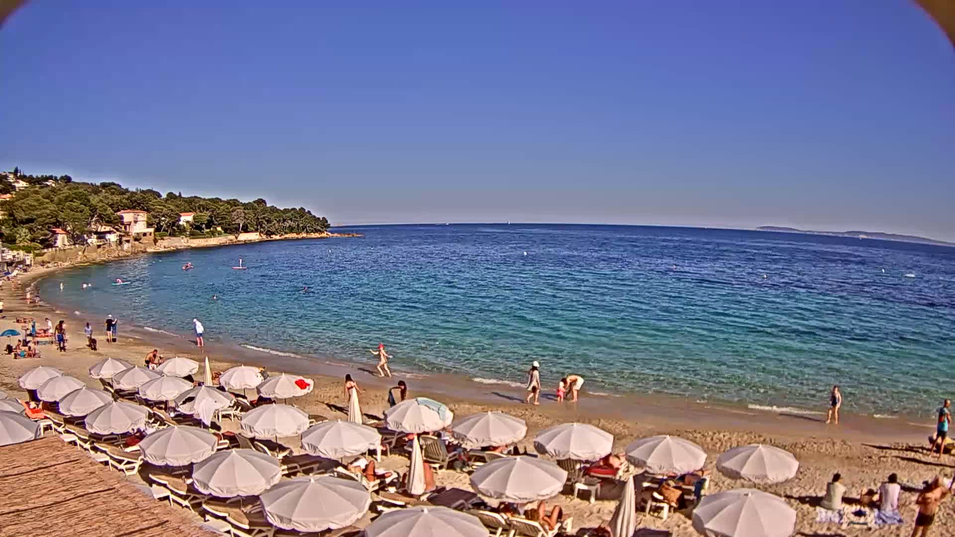 A sandy beach with numerous white umbrellas and sunbathers is situated next to a calm, clear turquoise sea under a bright blue sky.