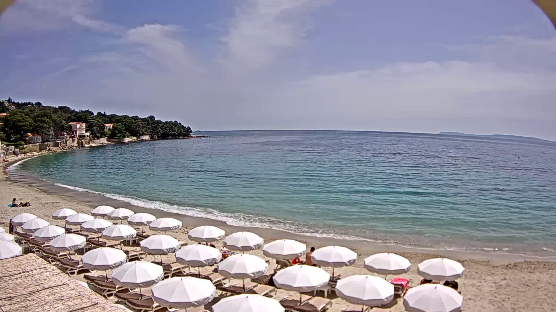 A sandy beach with numerous white umbrellas and lounge chairs is nestled in a calm, clear bay under a partly cloudy sky.