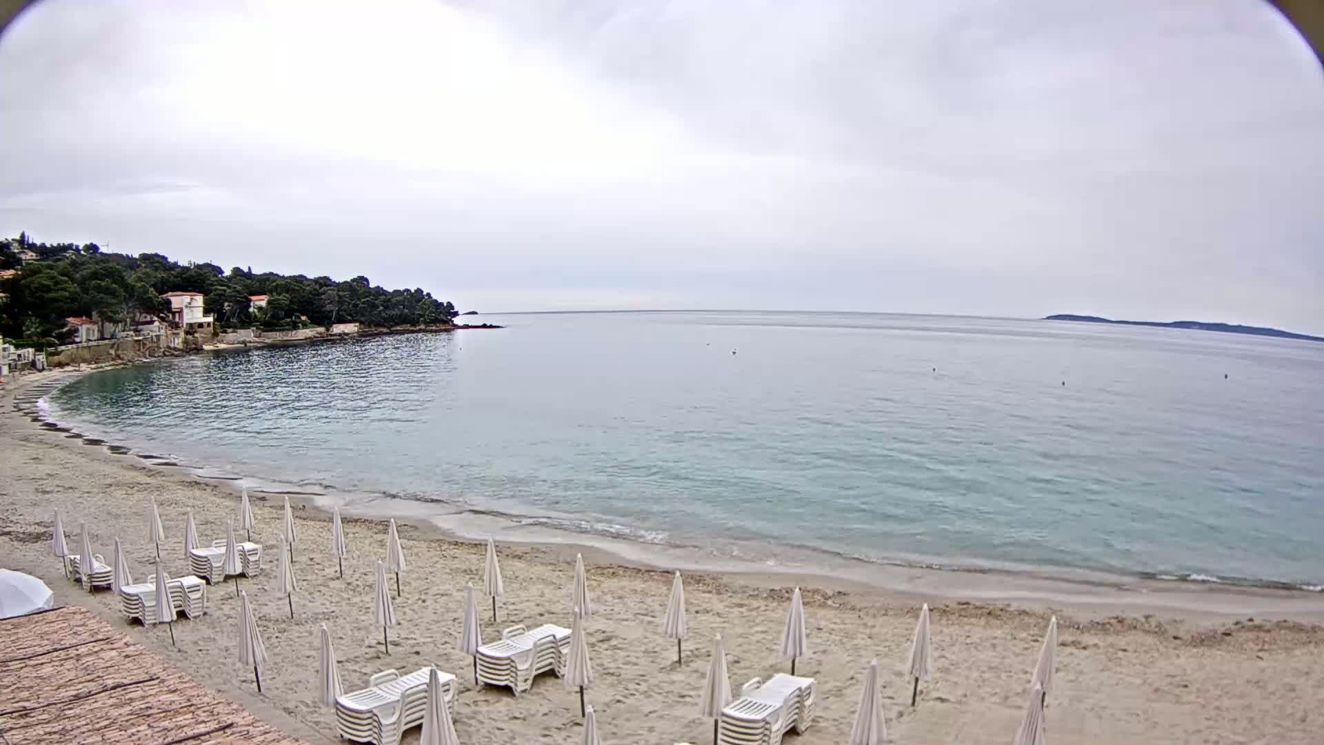 A sandy beach with white lounge chairs and umbrellas sits on a calm, light-blue sea under an overcast sky.
