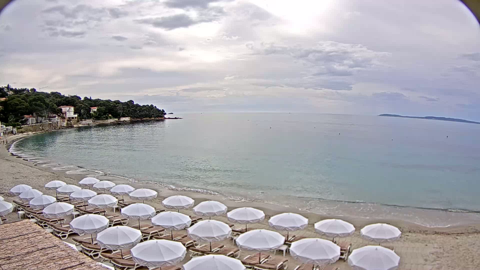 A sandy beach with rows of white umbrellas and lounge chairs is situated next to calm, light-blue water under a cloudy sky.