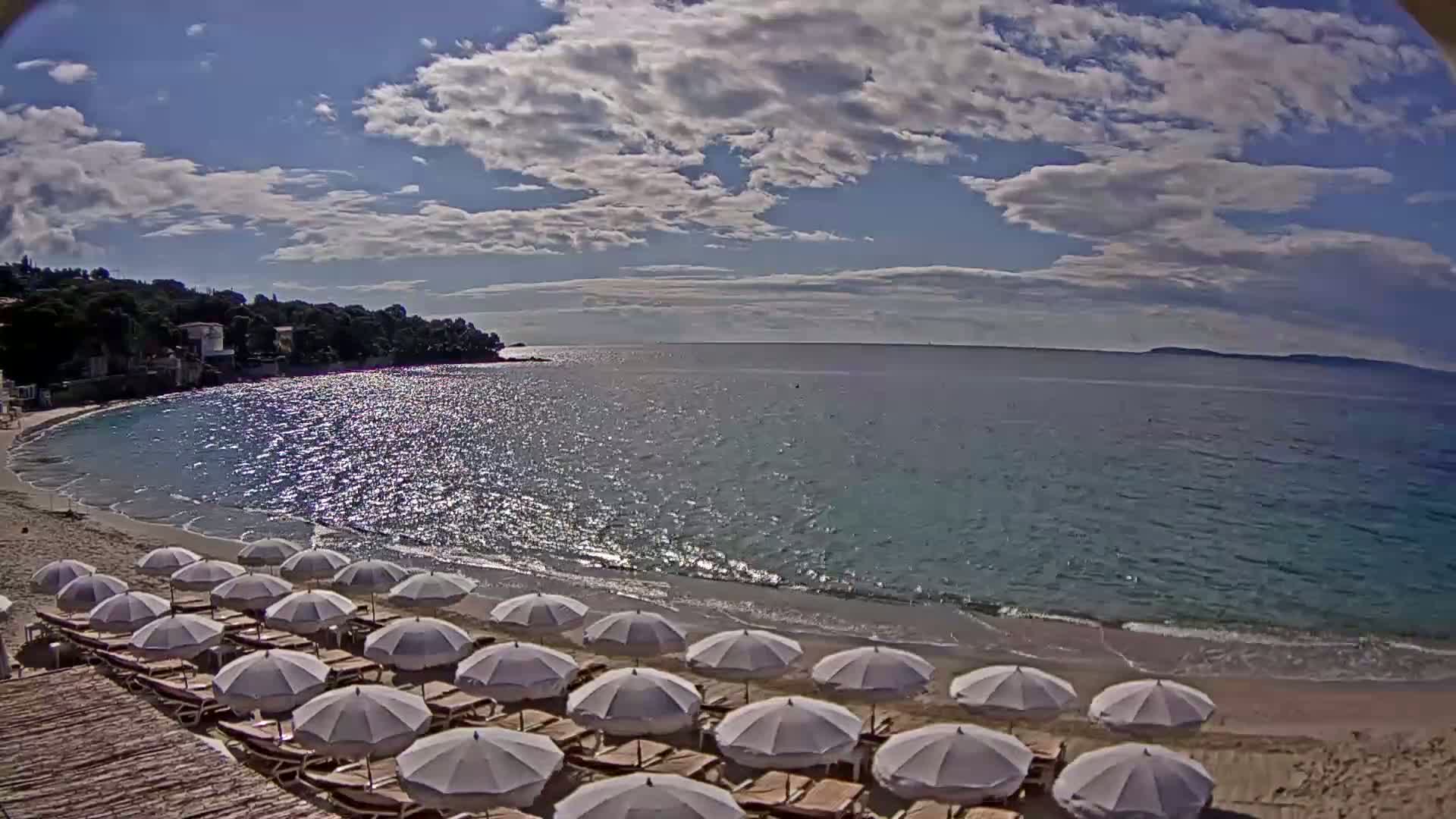 A sandy beach with rows of white umbrellas and lounge chairs is situated next to a calm, sparkling sea under a partly cloudy sky.