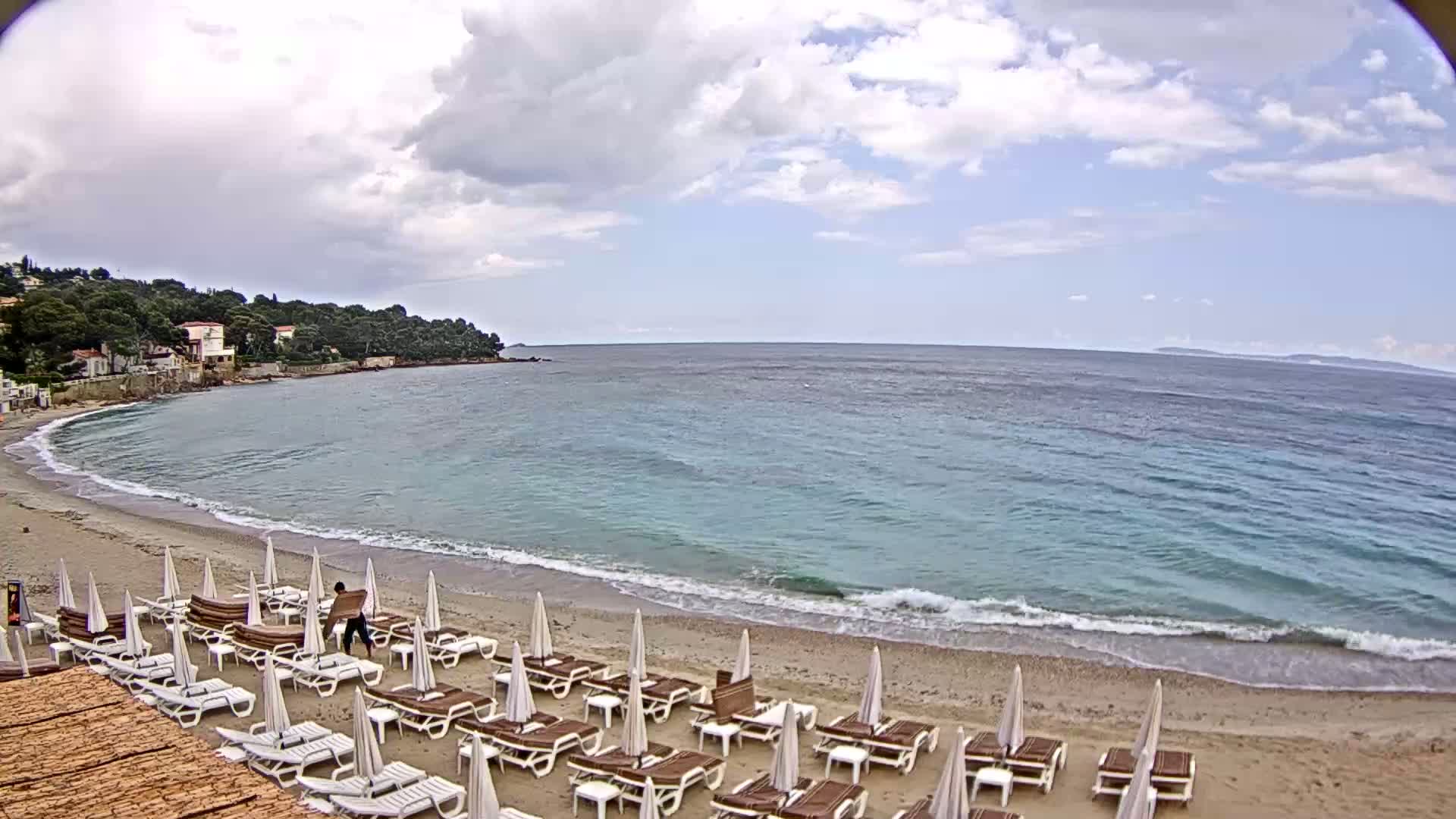 A sandy beach with rows of lounge chairs and umbrellas is partially obscured by a gently lapping sea under a partly cloudy sky.