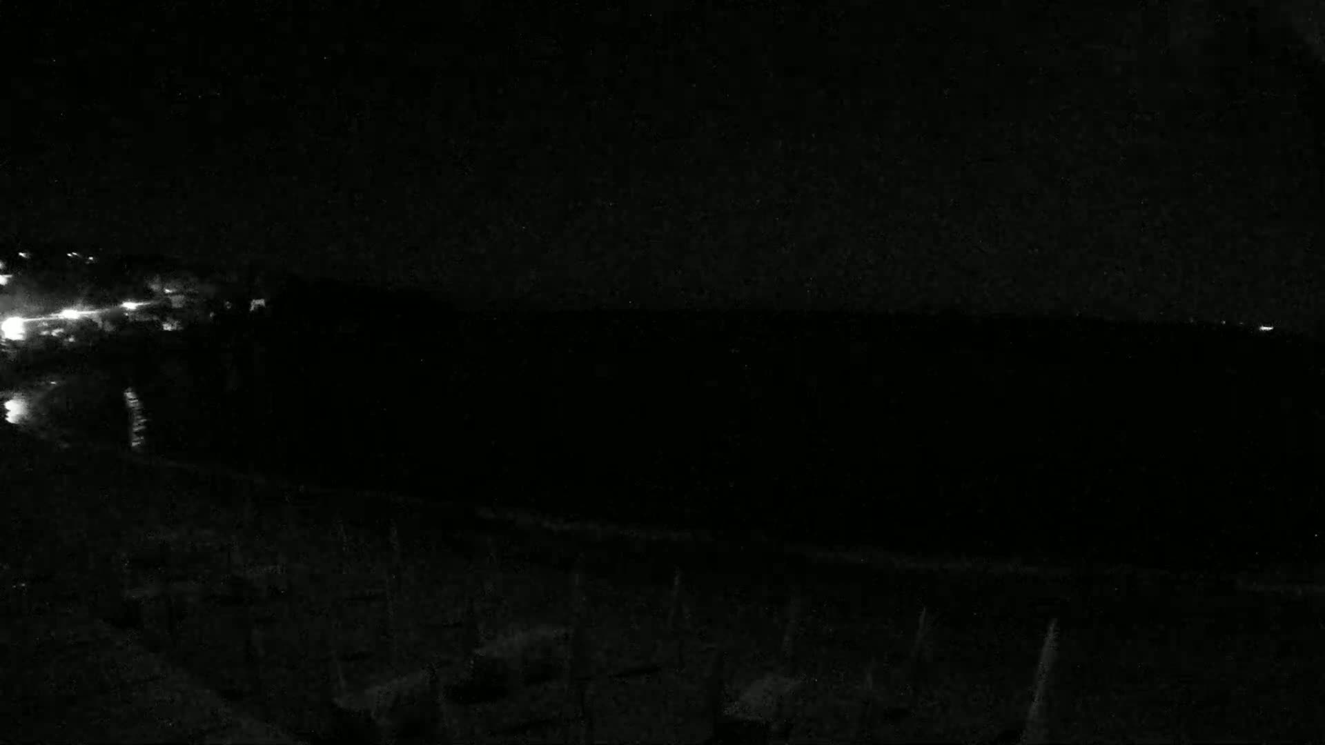 A nighttime, low-light view shows a dark body of water with a shoreline featuring sparse vegetation and faint lights in the distance.