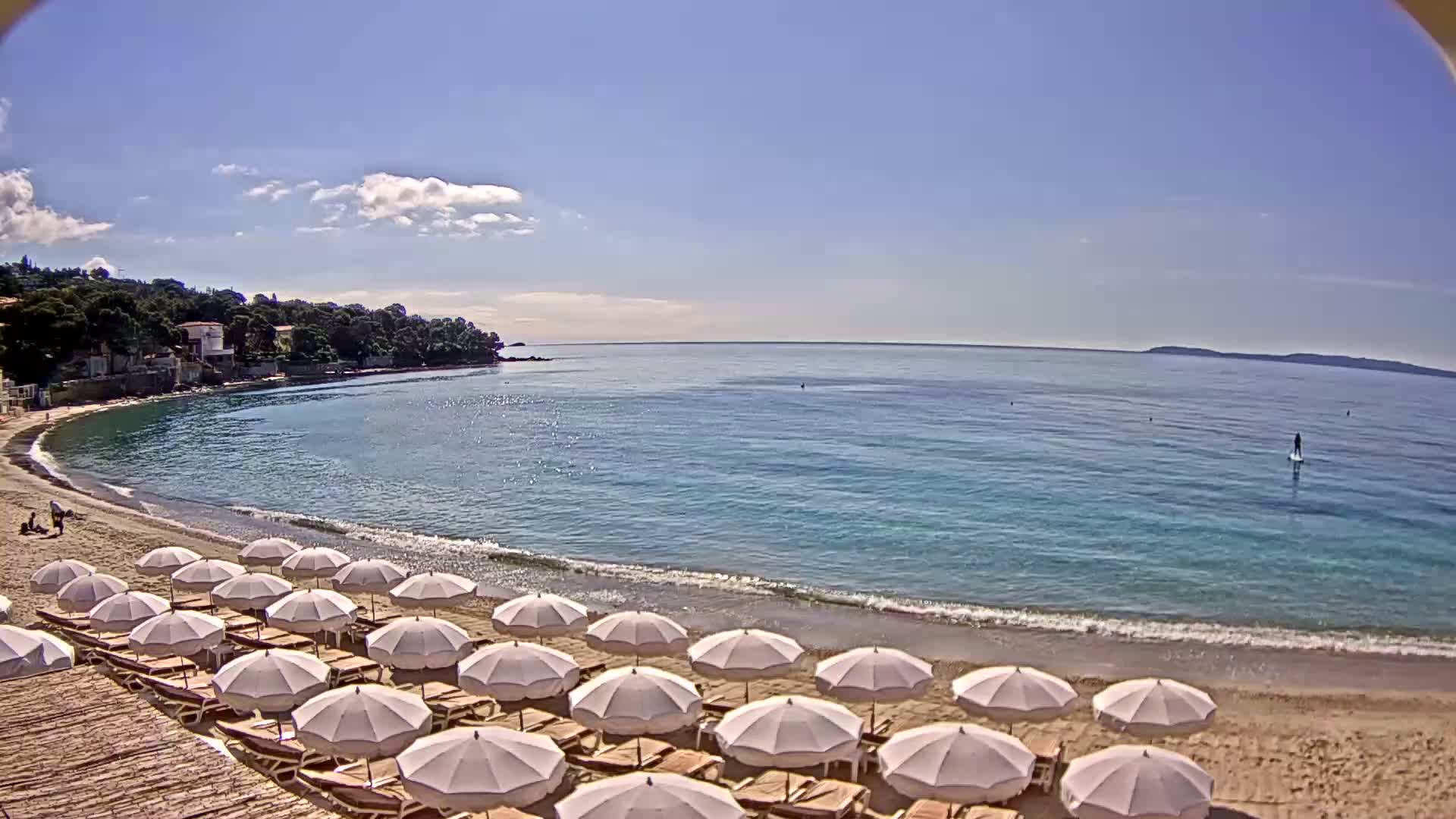 A sandy beach with rows of white umbrellas and lounge chairs is situated on a calm, clear blue sea under a mostly sunny sky.