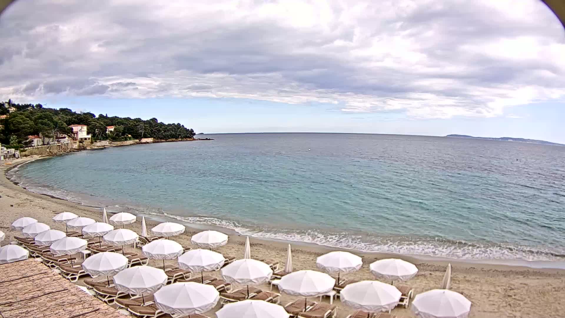 A sandy beach with numerous white umbrellas and lounge chairs is situated next to a calm, teal-colored sea under a partly cloudy sky.