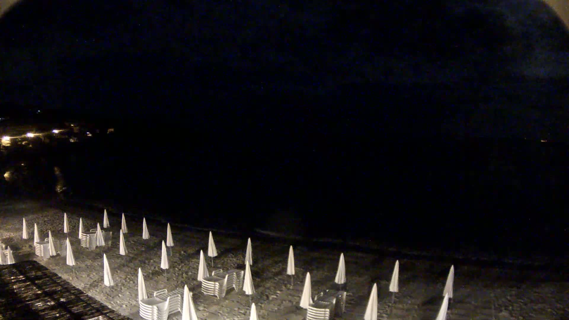 A dark beach at night is lined with closed beach umbrellas and chairs under a mostly clear sky.