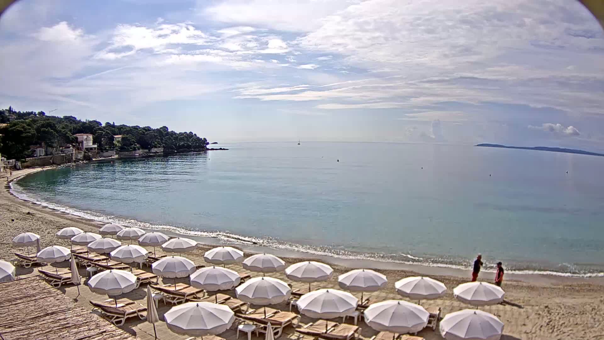 A sandy beach with rows of white umbrellas and lounge chairs overlooks a calm, light-blue sea under a partly cloudy sky.