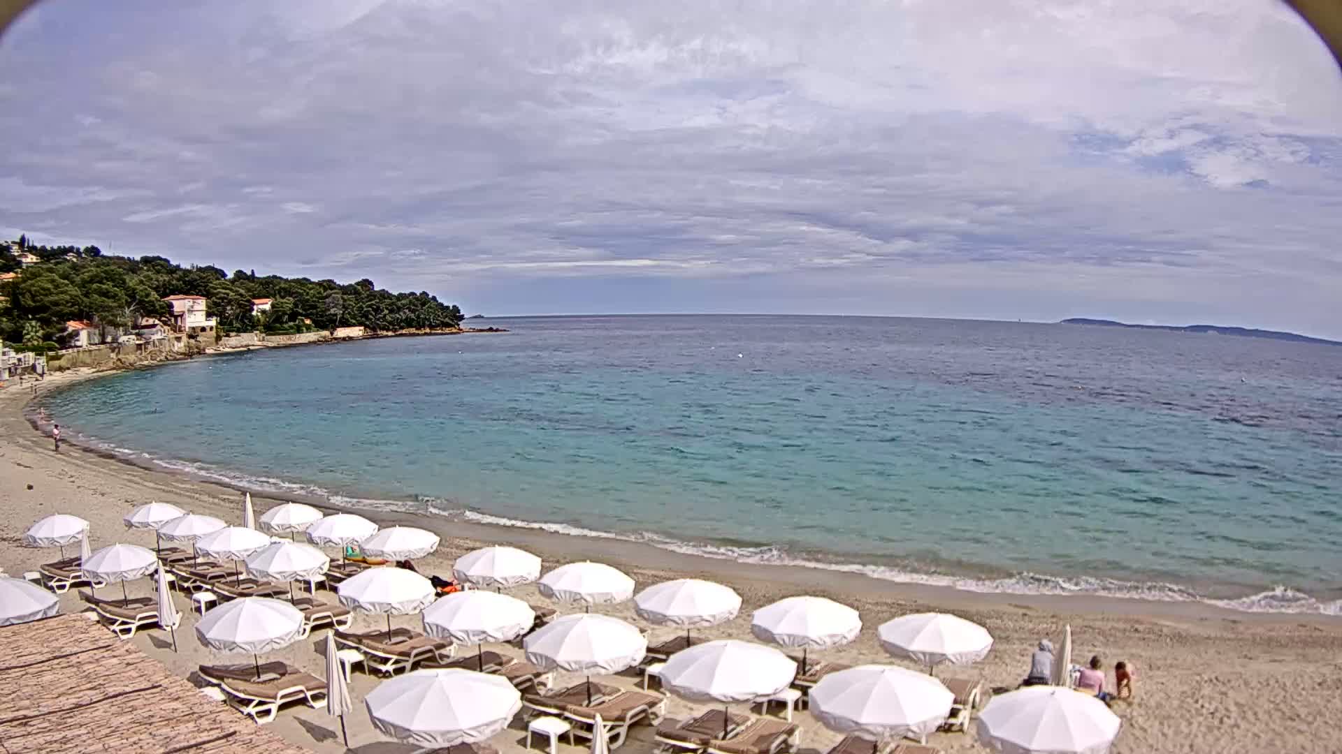 A sandy beach with rows of white umbrellas and lounge chairs is nestled in a cove of turquoise water under a partly cloudy sky.