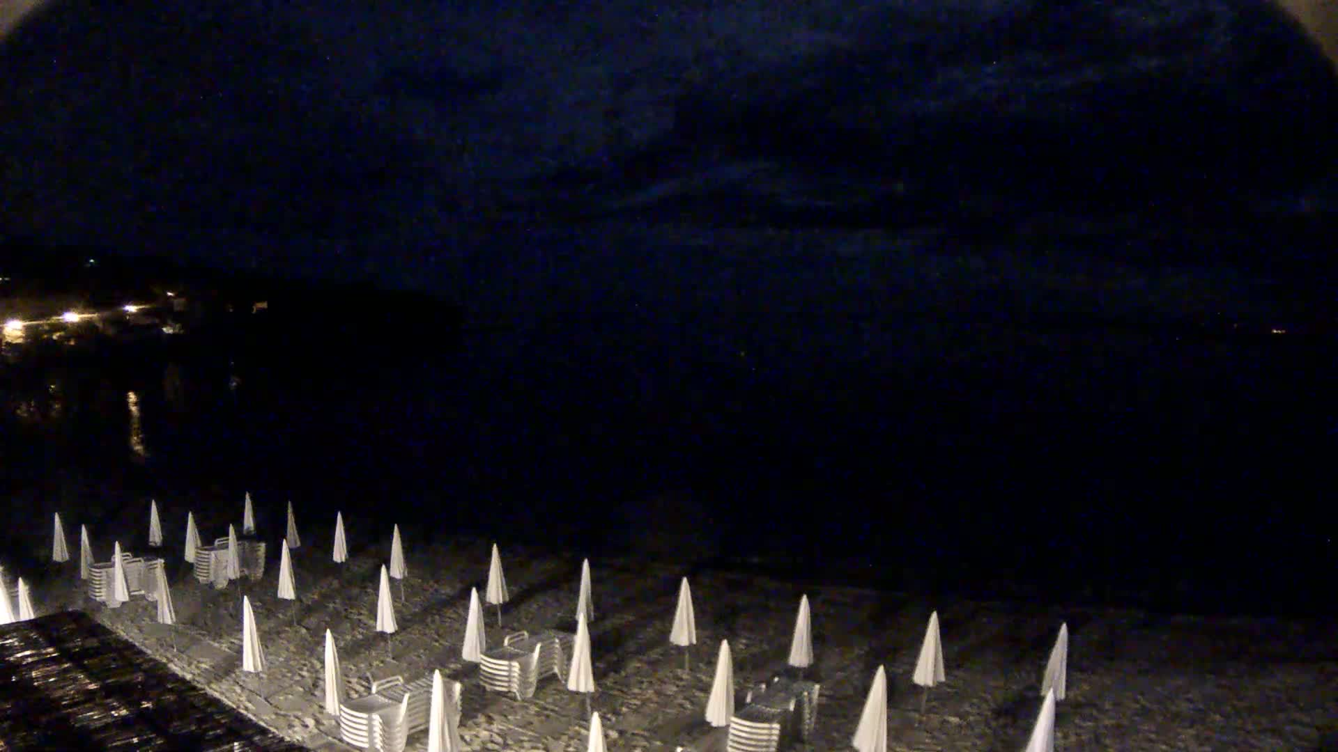 A dark beach at night, under a mostly cloudy sky, is lined with closed beach umbrellas and chairs.