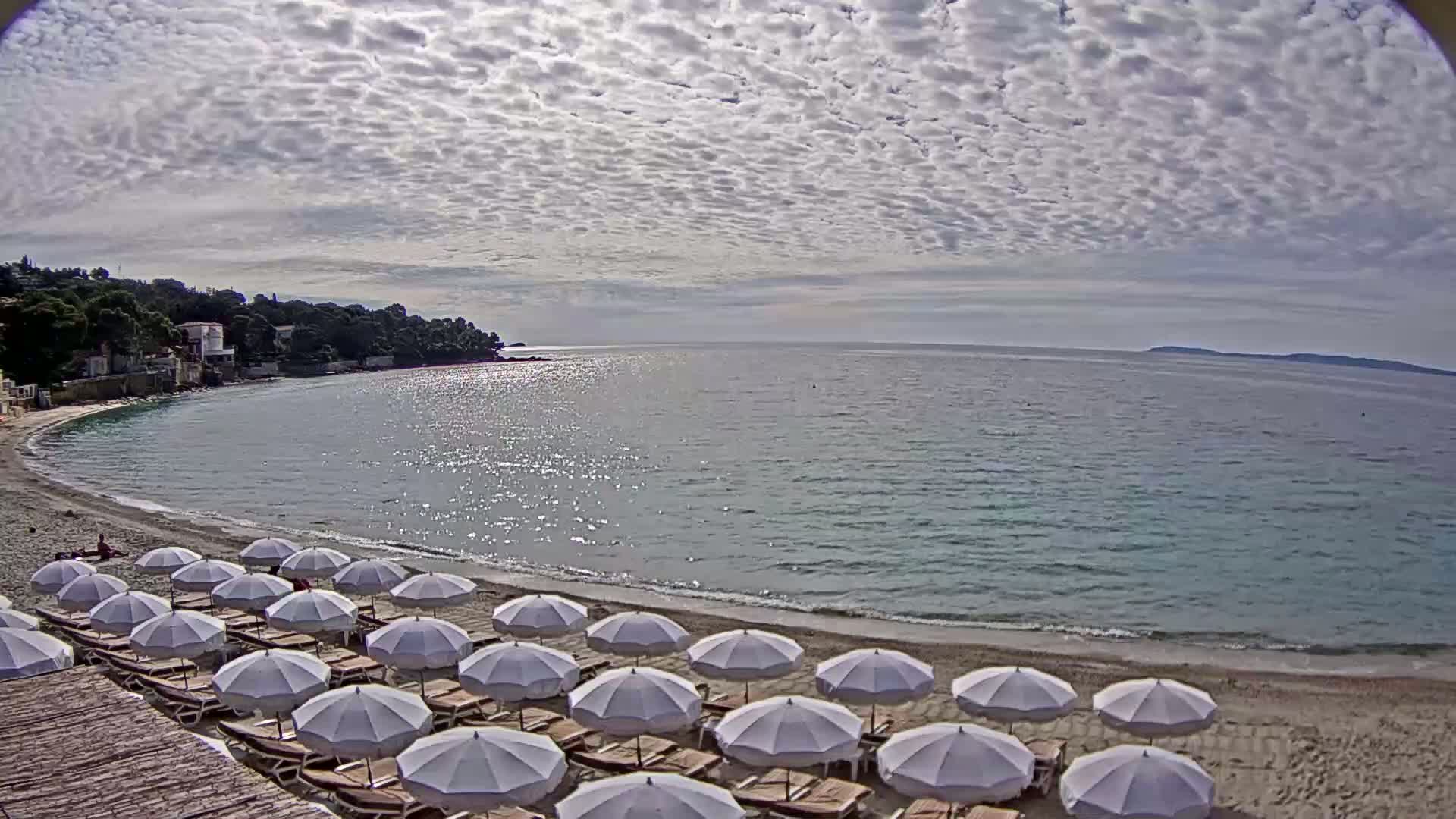 A sandy beach with rows of white umbrellas lines a calm, sparkling sea under a mostly cloudy sky.