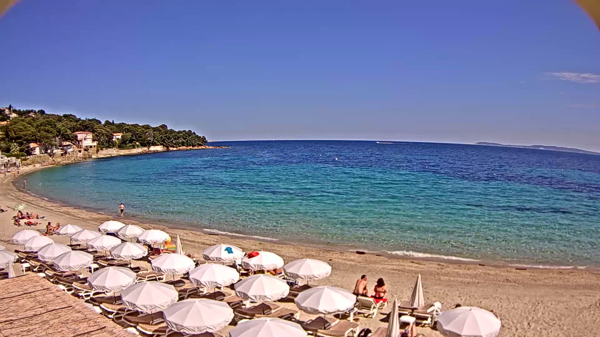 A sandy beach with white umbrellas and lounge chairs sits on a calm, clear turquoise ocean under a bright blue sky.