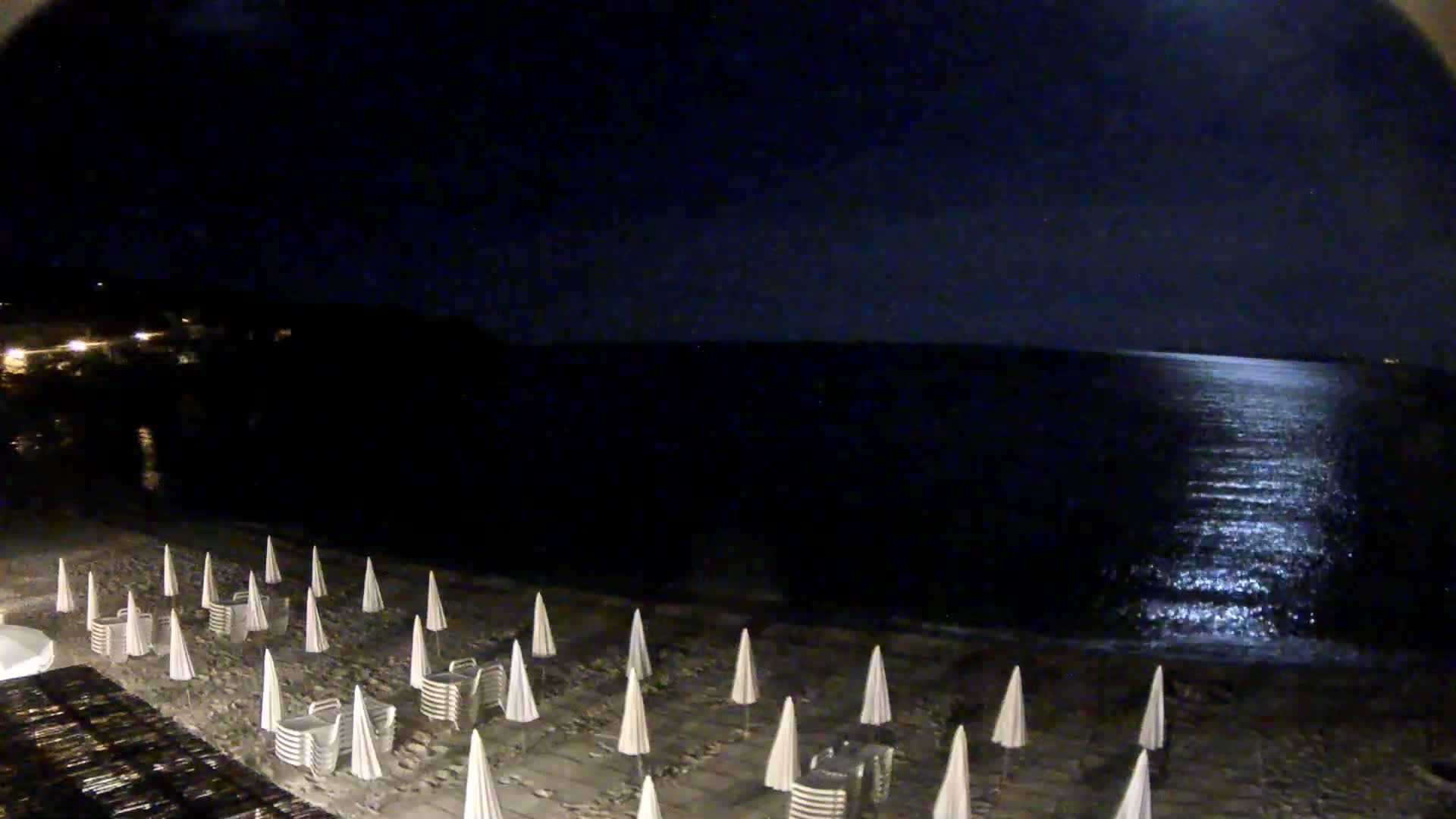 A moonlit beach at night shows rows of closed beach umbrellas on the sand, with a calm sea reflecting the light.