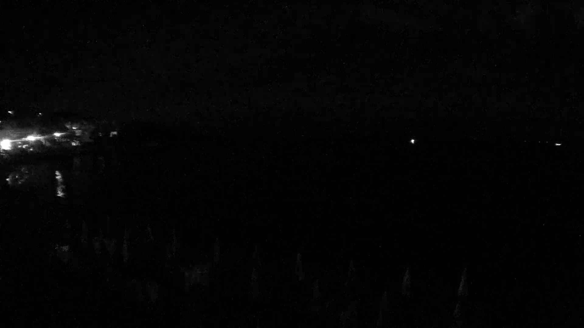 A nighttime view shows a dark landscape with a few distant lights and a dark body of water reflecting some light in the upper left.