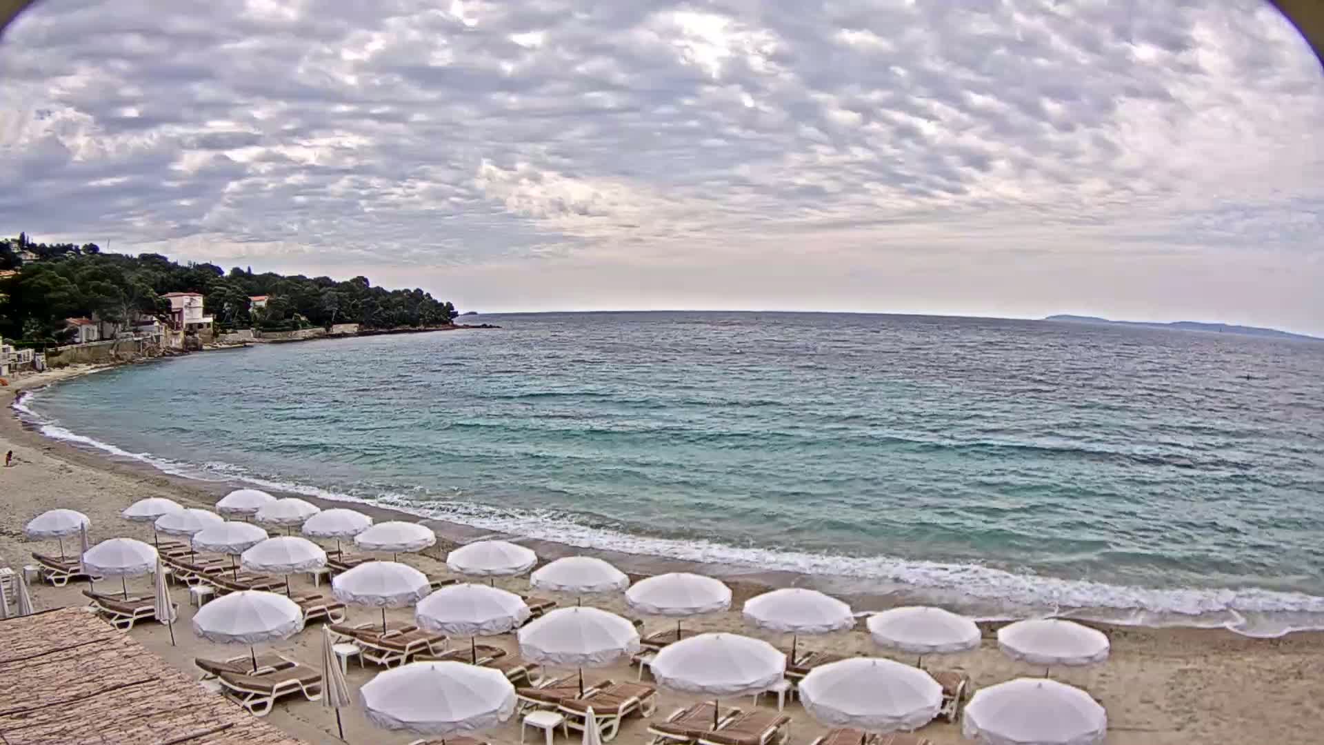 A sandy beach with rows of white umbrellas is situated next to a calm, turquoise sea under a cloudy sky.