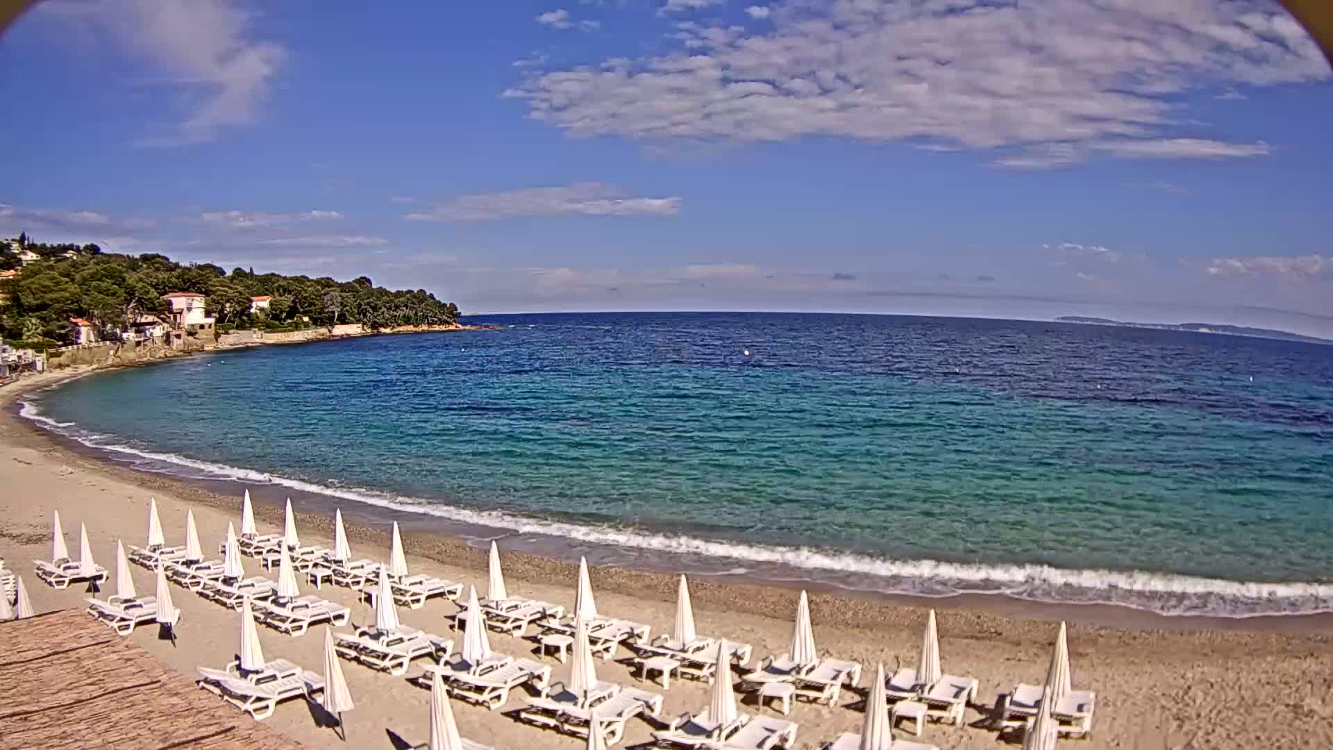 A sandy beach with rows of white lounge chairs and umbrellas under a partly cloudy blue sky, borders a calm, turquoise sea.