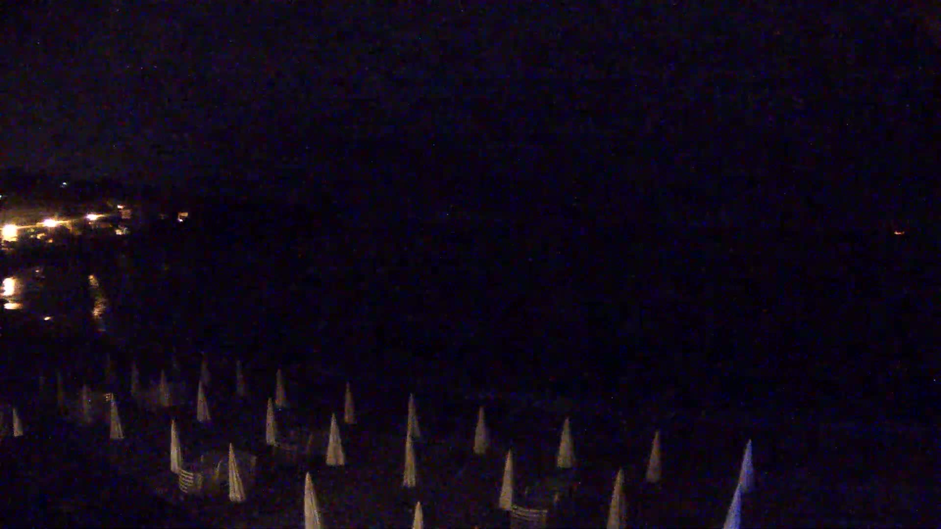 At night, a dark beach with many closed beach umbrellas is visible, and a body of water with distant lights reflects light.