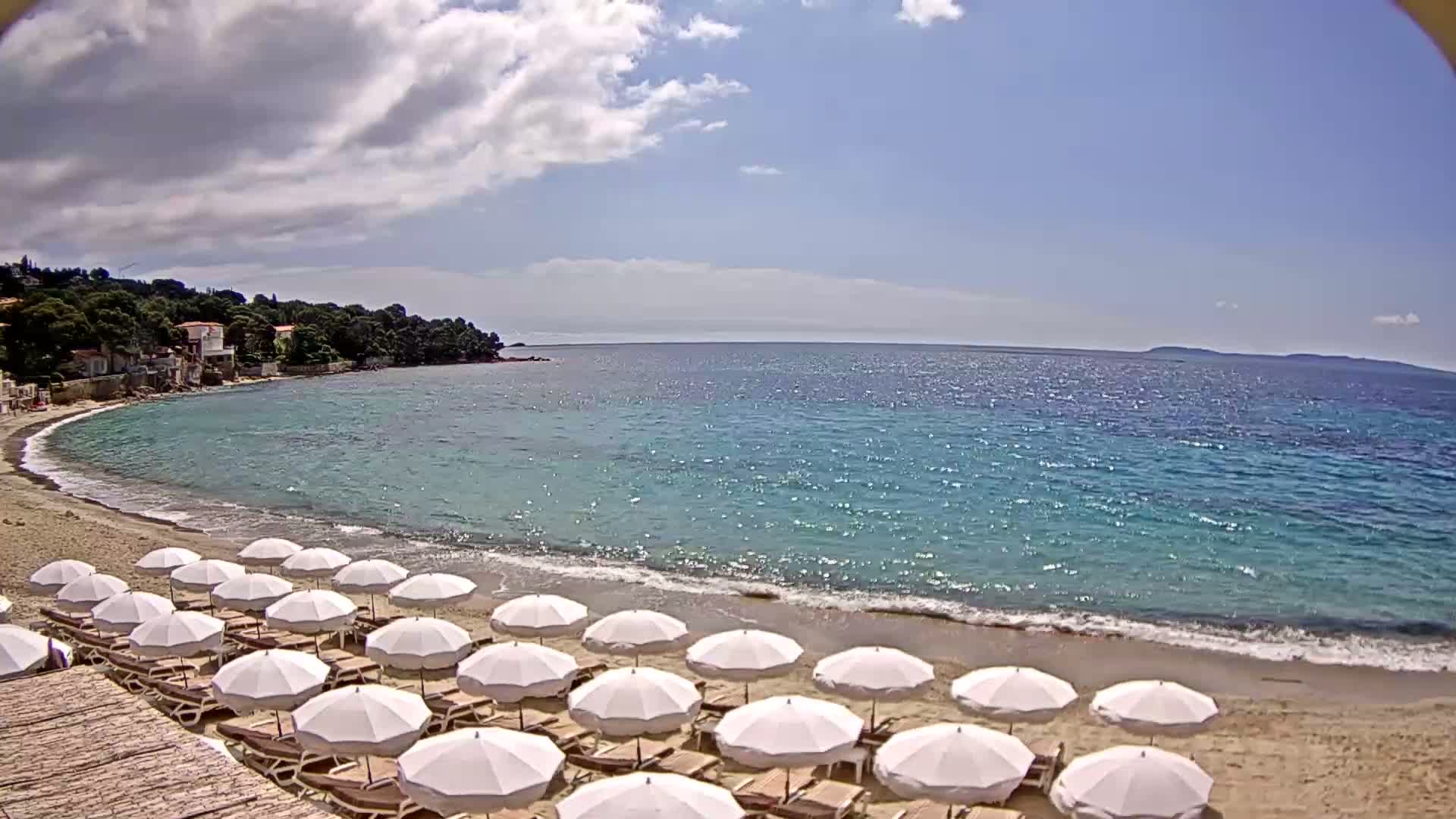 A sandy beach with rows of white umbrellas and lounge chairs lines a calm, clear turquoise sea under a partly cloudy sky.