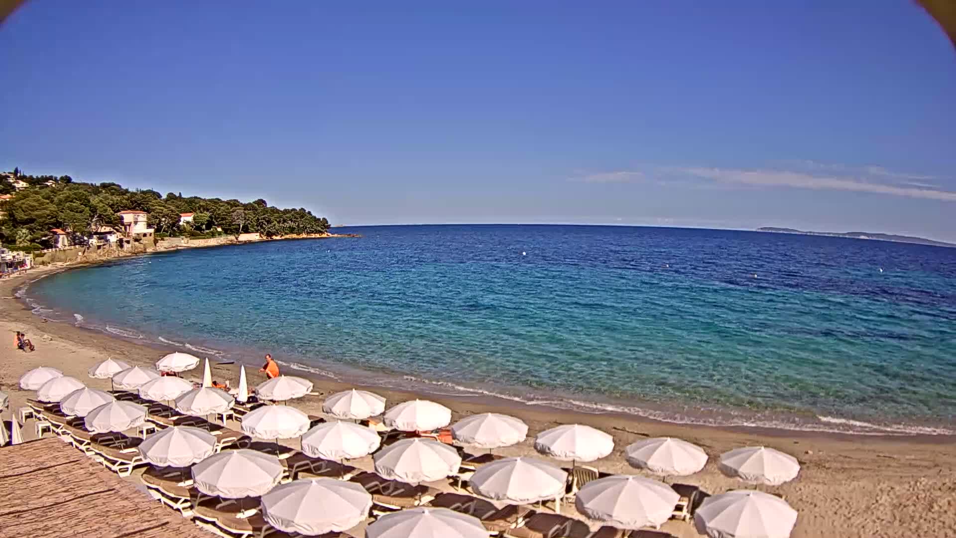 A sandy beach with numerous white umbrellas and lounge chairs lines a calm, clear turquoise ocean under a bright blue sky.