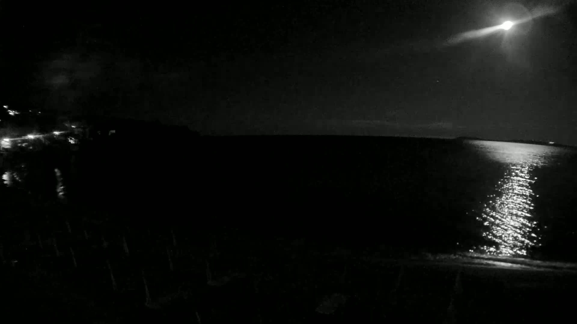 A bright moon illuminates a dark, calm body of water at night, with a shoreline visible in the foreground.