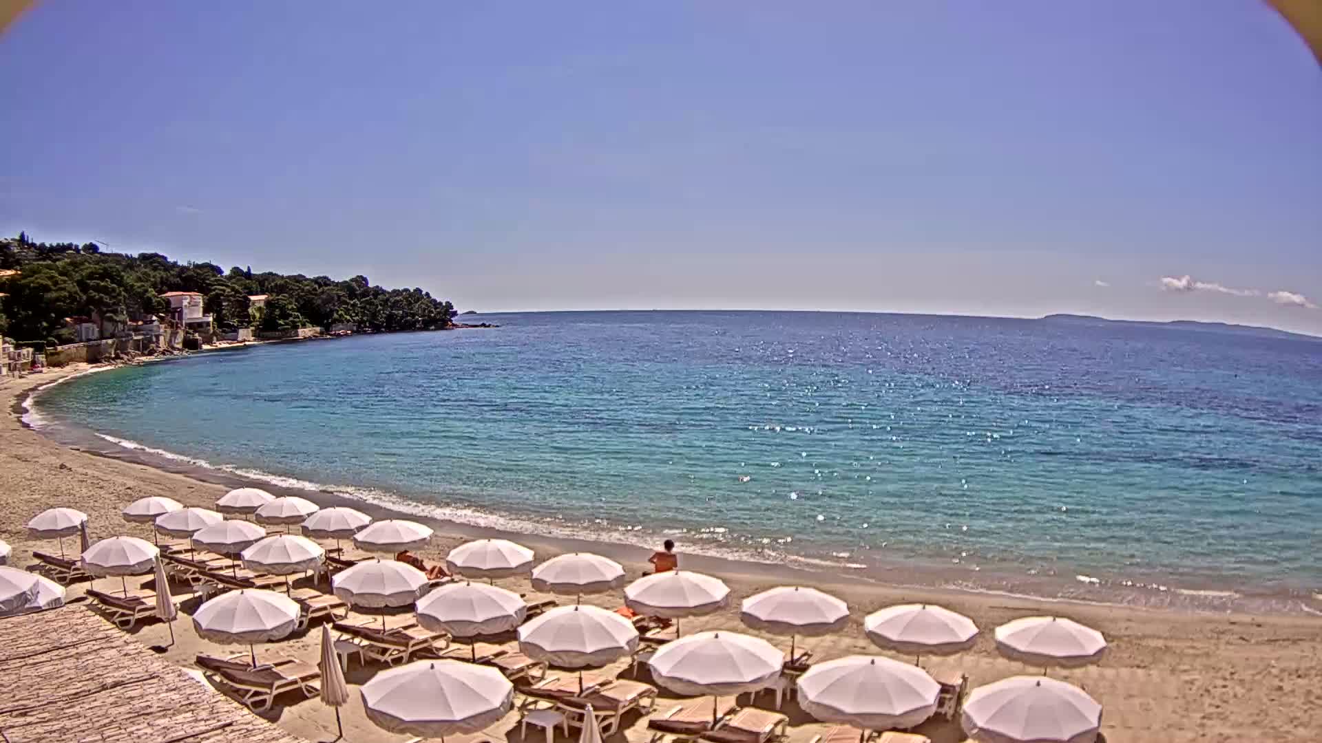 A sandy beach with numerous white umbrellas and lounge chairs lines a calm, clear turquoise sea under a bright sunny sky, backed by a small coastal town and green hills.