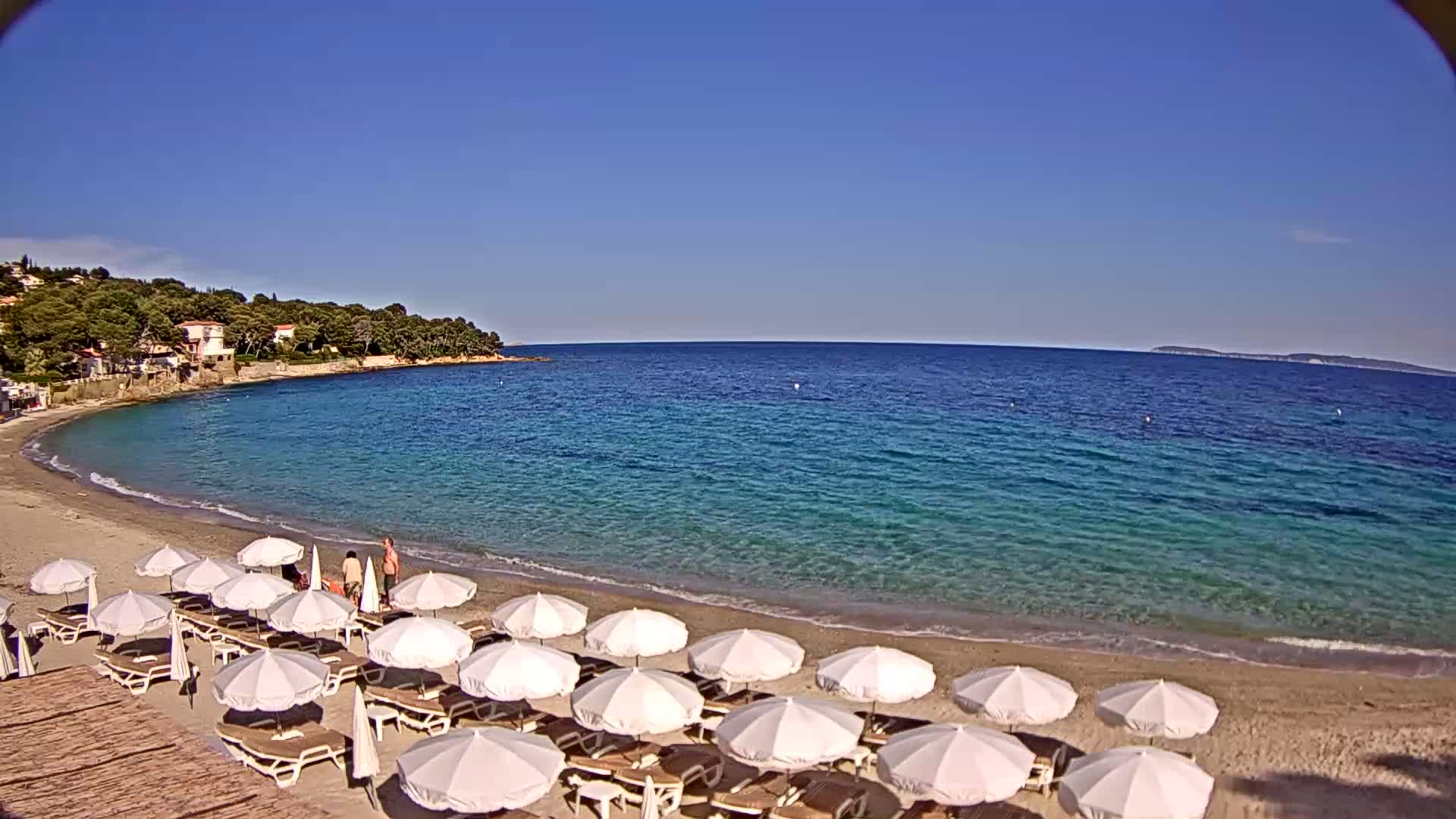A sandy beach with white umbrellas and lounge chairs lines a calm, clear turquoise sea under a bright blue sky.