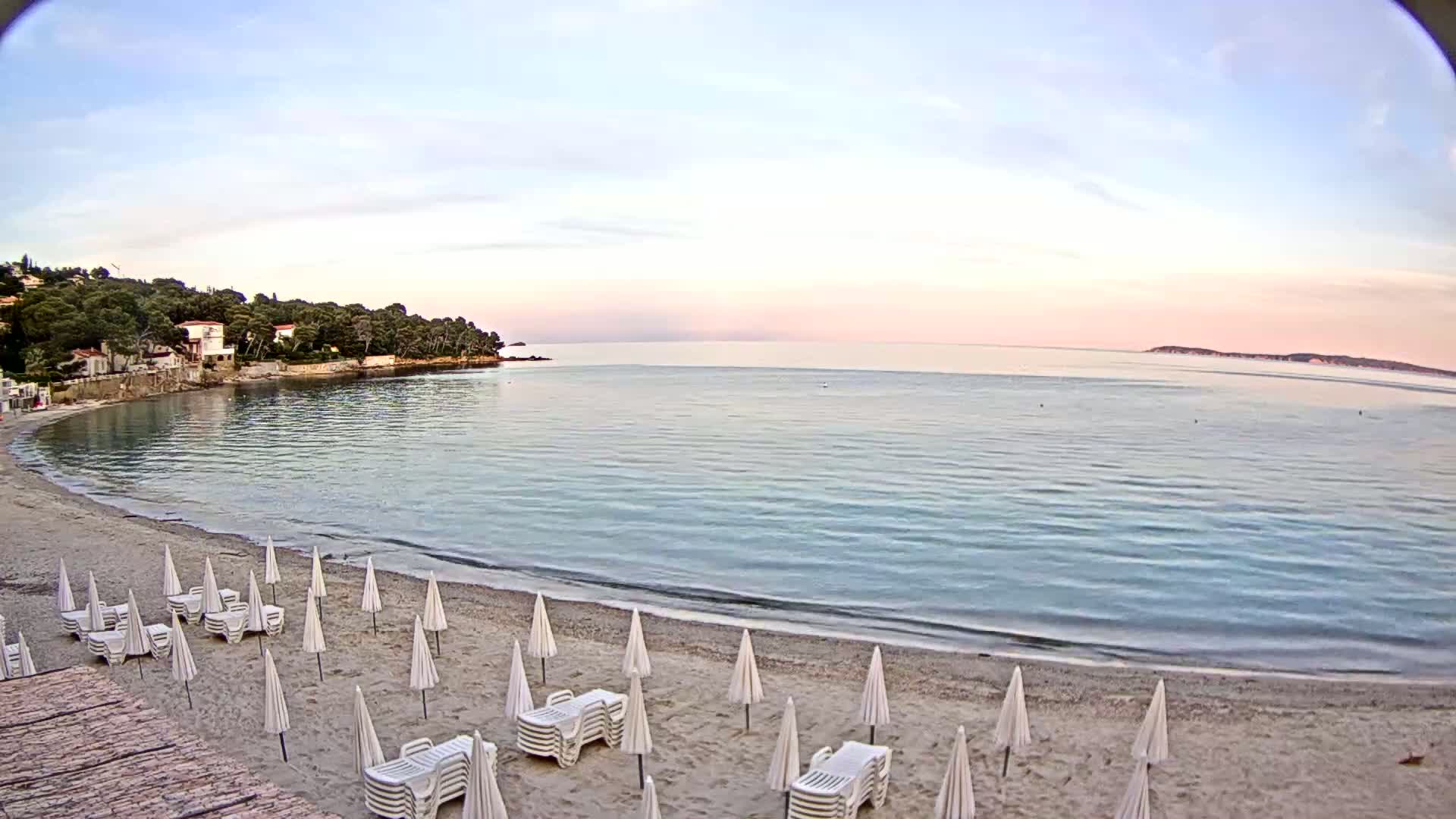A calm, pale-pink-hued beach with white umbrellas and lounge chairs sits at the edge of a tranquil bay under a clear sky.