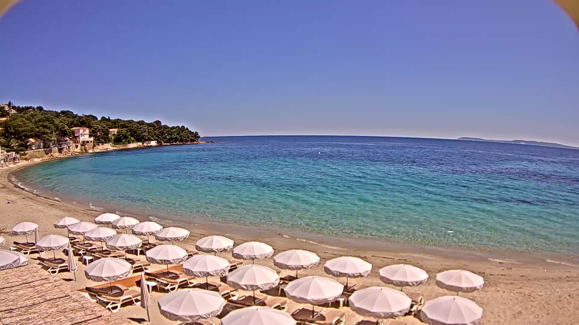 A sandy beach with rows of white umbrellas and lounge chairs curves around a calm, clear turquoise sea under a bright blue sky.