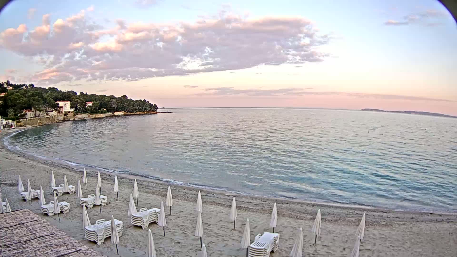 A sandy beach with white lounge chairs and umbrellas sits under a partly cloudy, pinkish sunset next to a calm sea and a tree-lined shore.