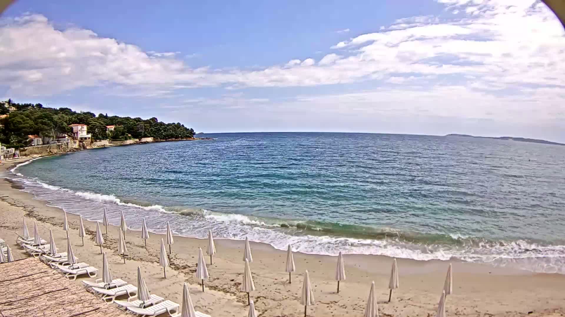 A sandy beach with white umbrellas and lounge chairs is bordered by a calm, blue sea under a partly cloudy sky.