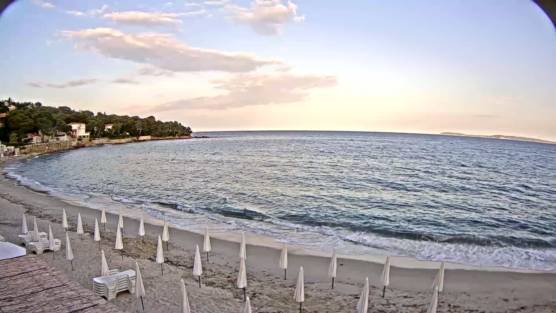 A sandy beach with white umbrellas and lounge chairs is gently lapped by waves on a calm sea under a partly cloudy sky.