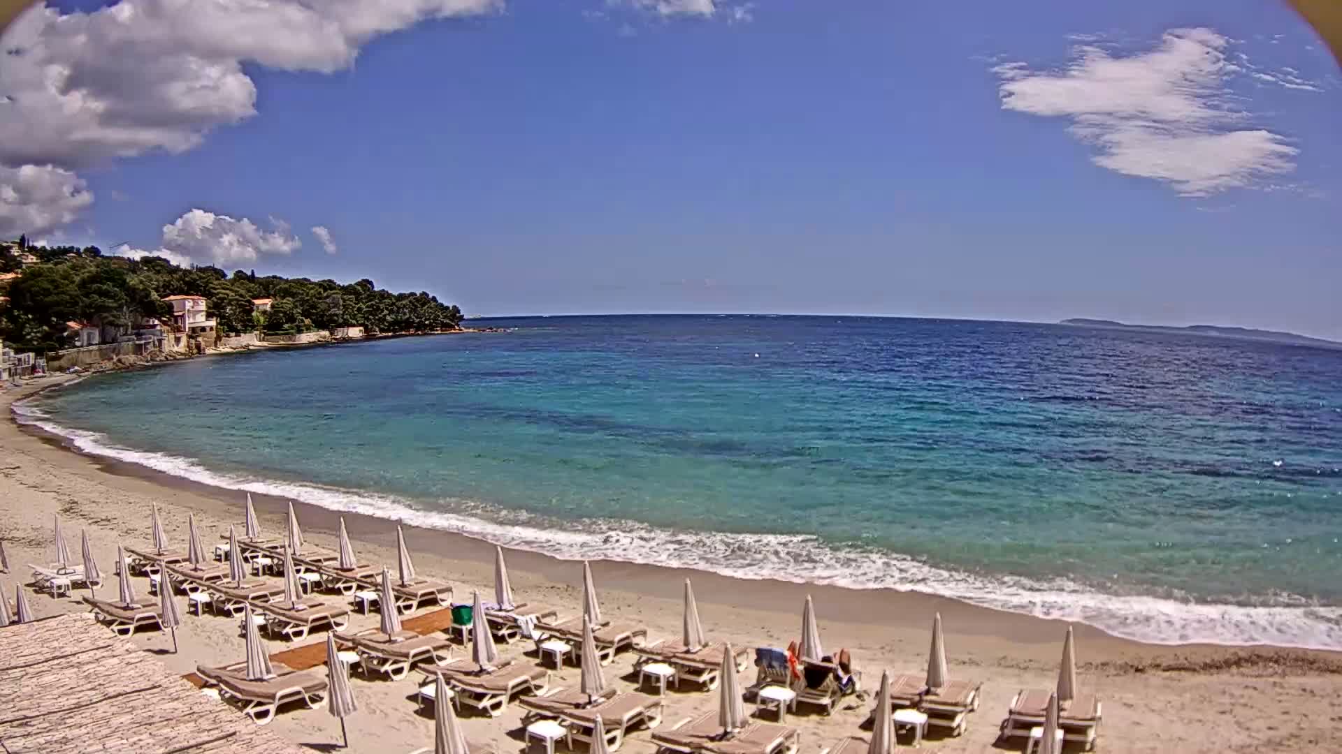 A sandy beach with rows of lounge chairs and umbrellas is situated next to a calm, teal-colored ocean under a partly cloudy sky.